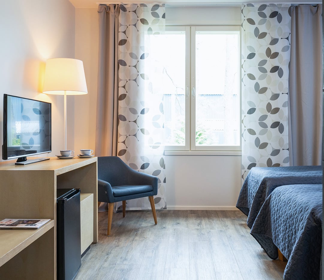Vaalea hotellihuone, aurinko paistaa huoneeseen, kaksi sänkyä pöytä, nojatuoli ja televisio