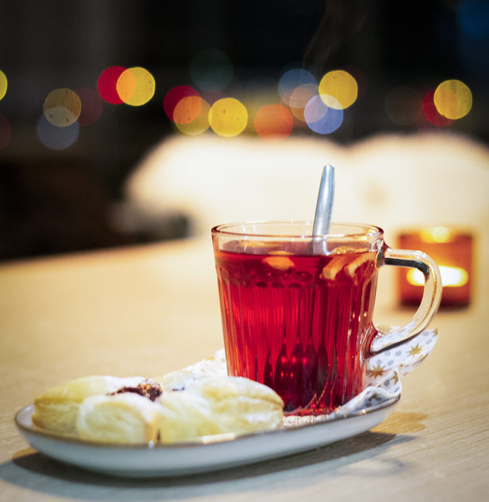 Glögi lasimukissa, joulutorttu lautasella ja kimmeltäviä valoja ja kynttilä