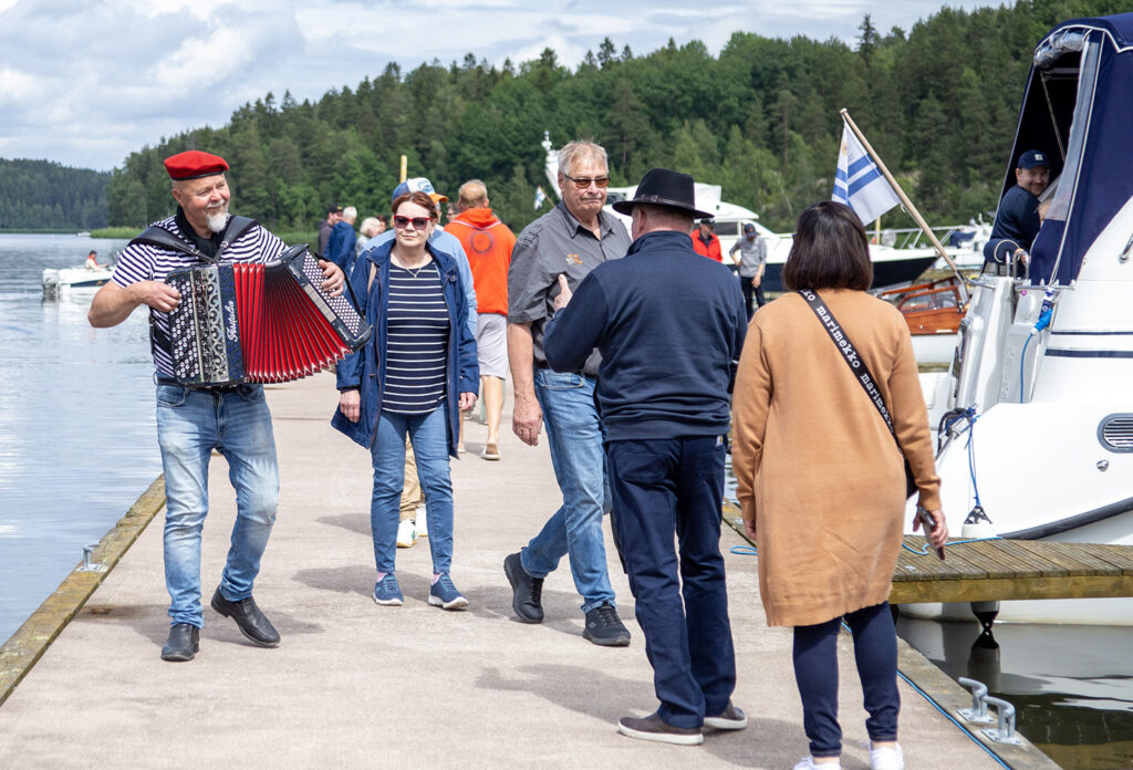 Iloisesti rankalaisittain punaiseen baskeriin ja raidalliseen paitaan pukeutunut haitarin soittaja kävelee satamassa laiturilla yleisön seassa